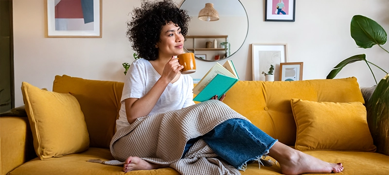 A woman smiles while relaxing on her couch reading a book and sipping from a mug, representing positive mental health in the new year