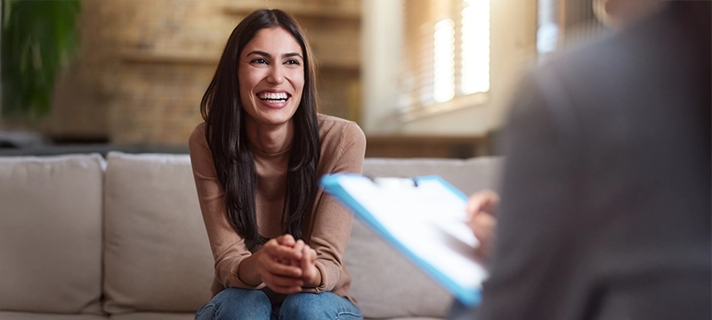 A brunette woman smiles brightly during a psychological evaluation with a clinical neuropsychologist sitting across from her, representative of using PAR products for positive patient outcomes